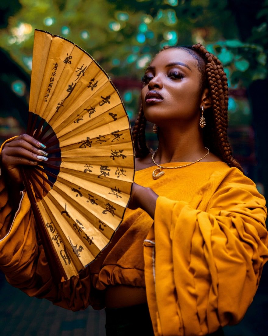 Woman with braids in a mustard outfit holding a gold fan
